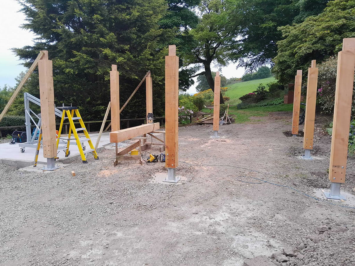 Timber posts erected on steel base plates at the start of car port construction, with yellow stepladder on site