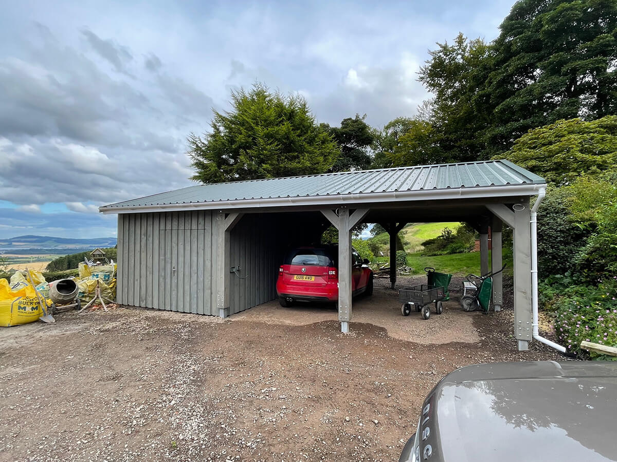Completed timber car port with grey standing seam roof and vertical cladding, red car parked inside on gravel drive