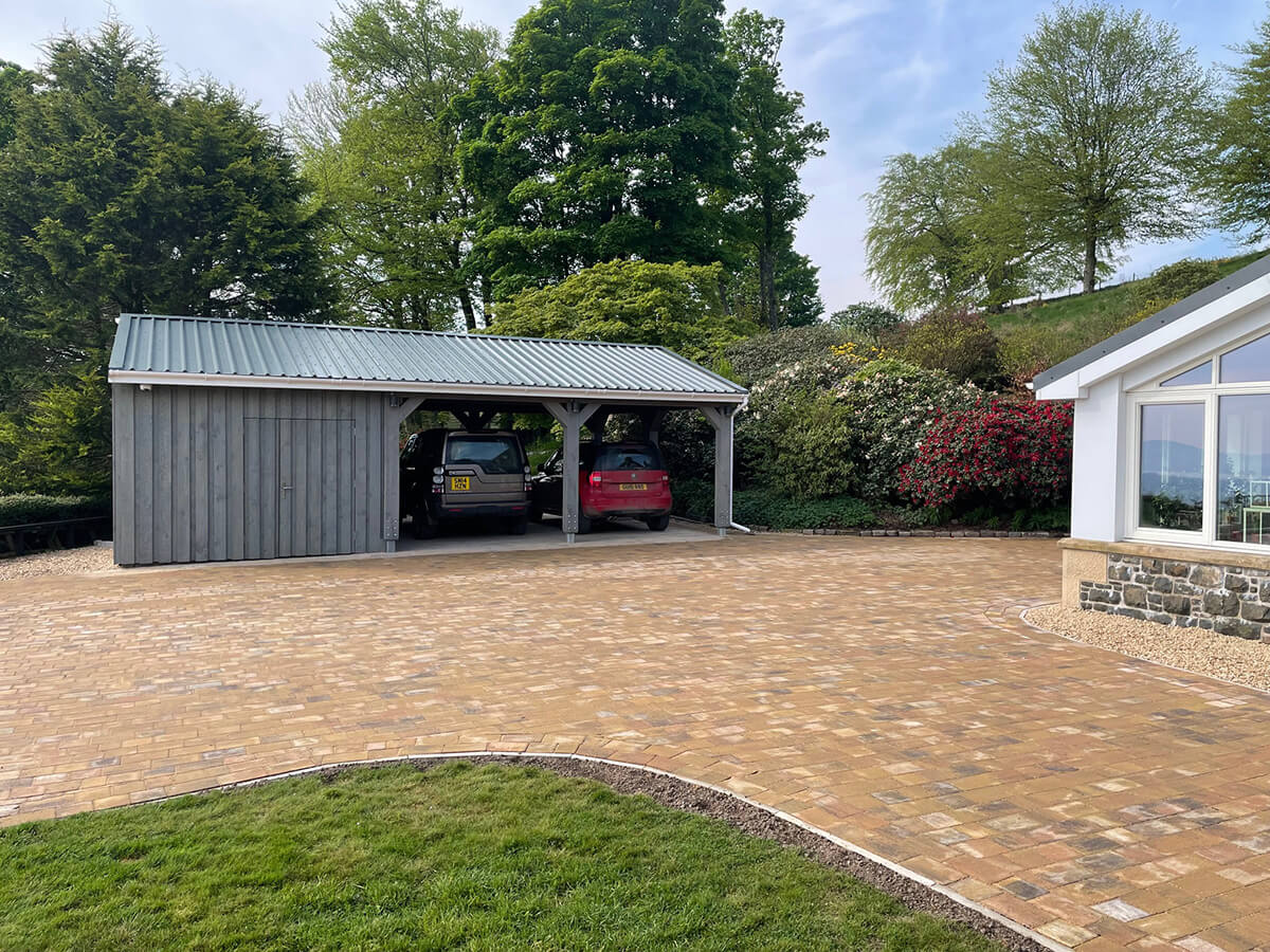 Completed timber car port with standing seam roof and grey cladding, two cars parked on block-paved driveway beside the house