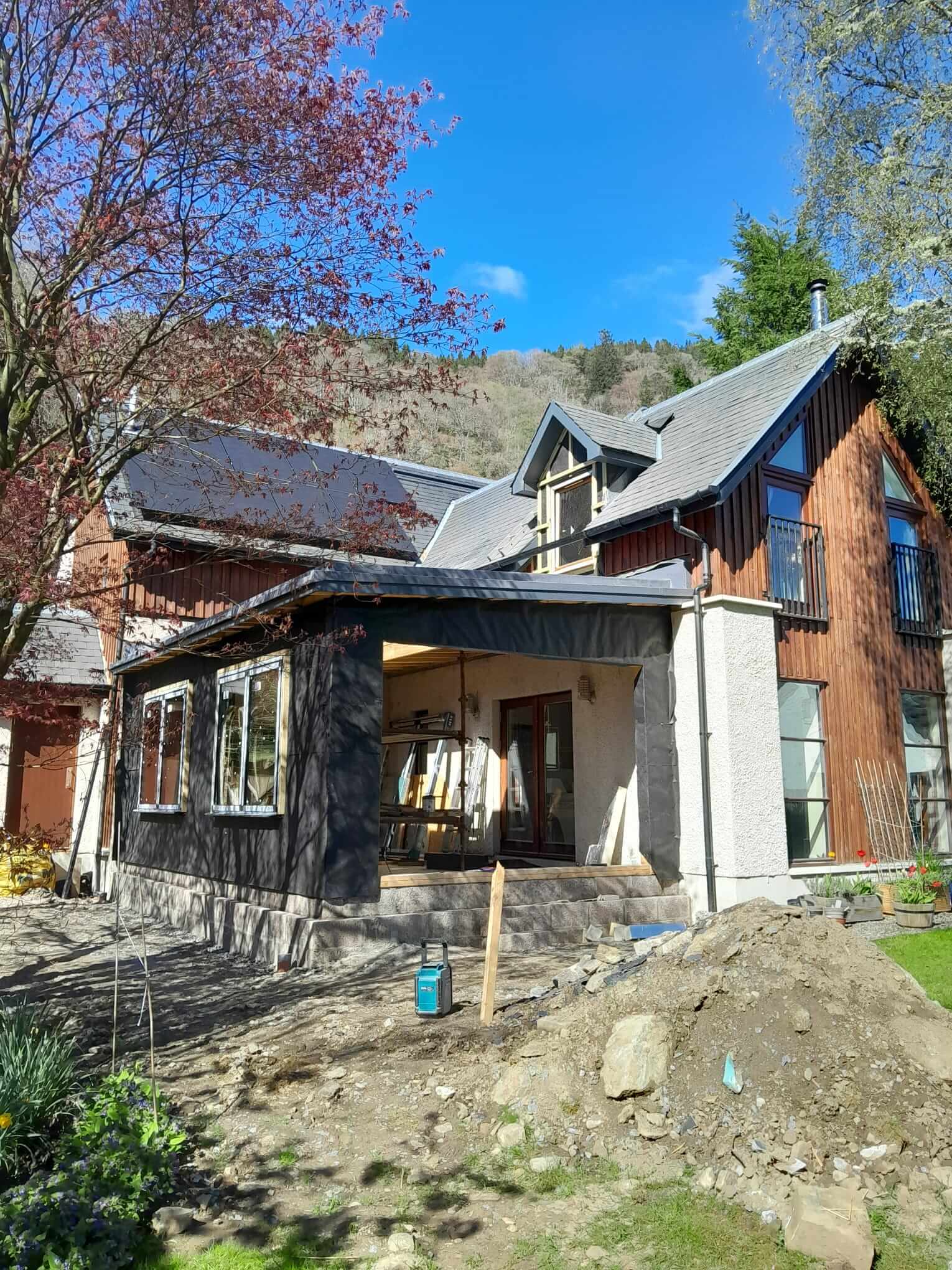 Near-complete extension exterior from garden, dark breather membrane applied, flat roof complete, anthracite glazing frames visible, red Japanese maple in foreground
