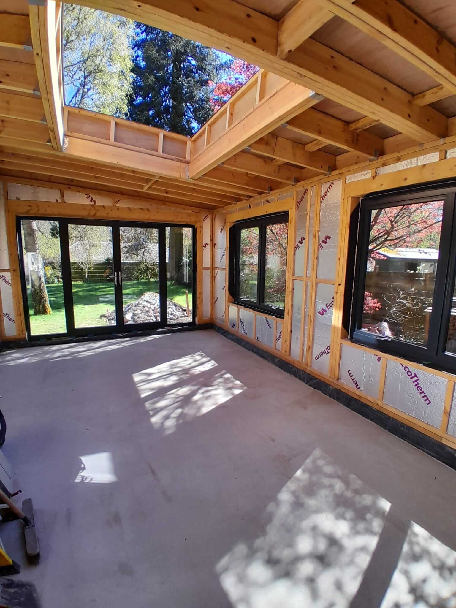 Interior of extension with Ecotherm insulation fitted between timber studs, anthracite aluminium bifold doors and corner window installed, bespoke rooflight open to sky above polished concrete screed