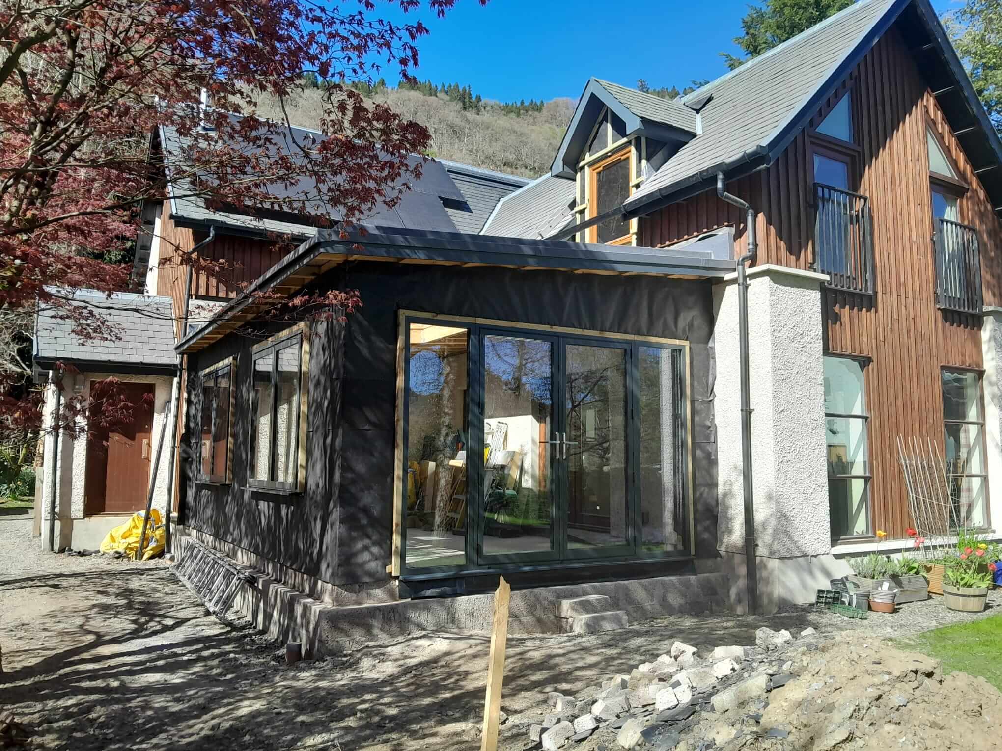 Exterior elevation showing anthracite aluminium bifold doors fitted to extension, dark breather membrane awaiting render, larch-clad main house behind