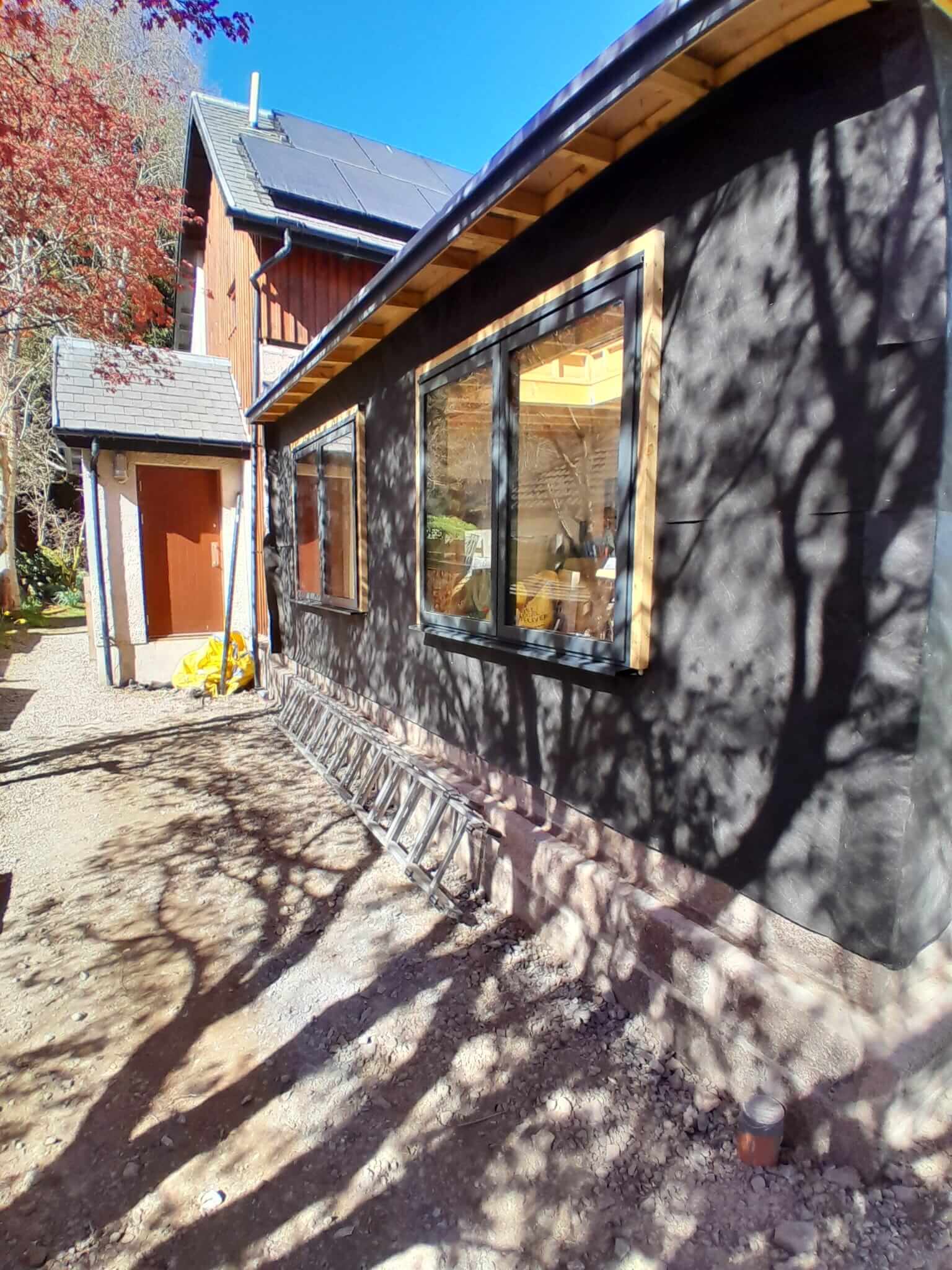 Side elevation of extension showing anthracite aluminium windows fitted, dark breather membrane with tree shadows cast across wall, larch-clad main house behind