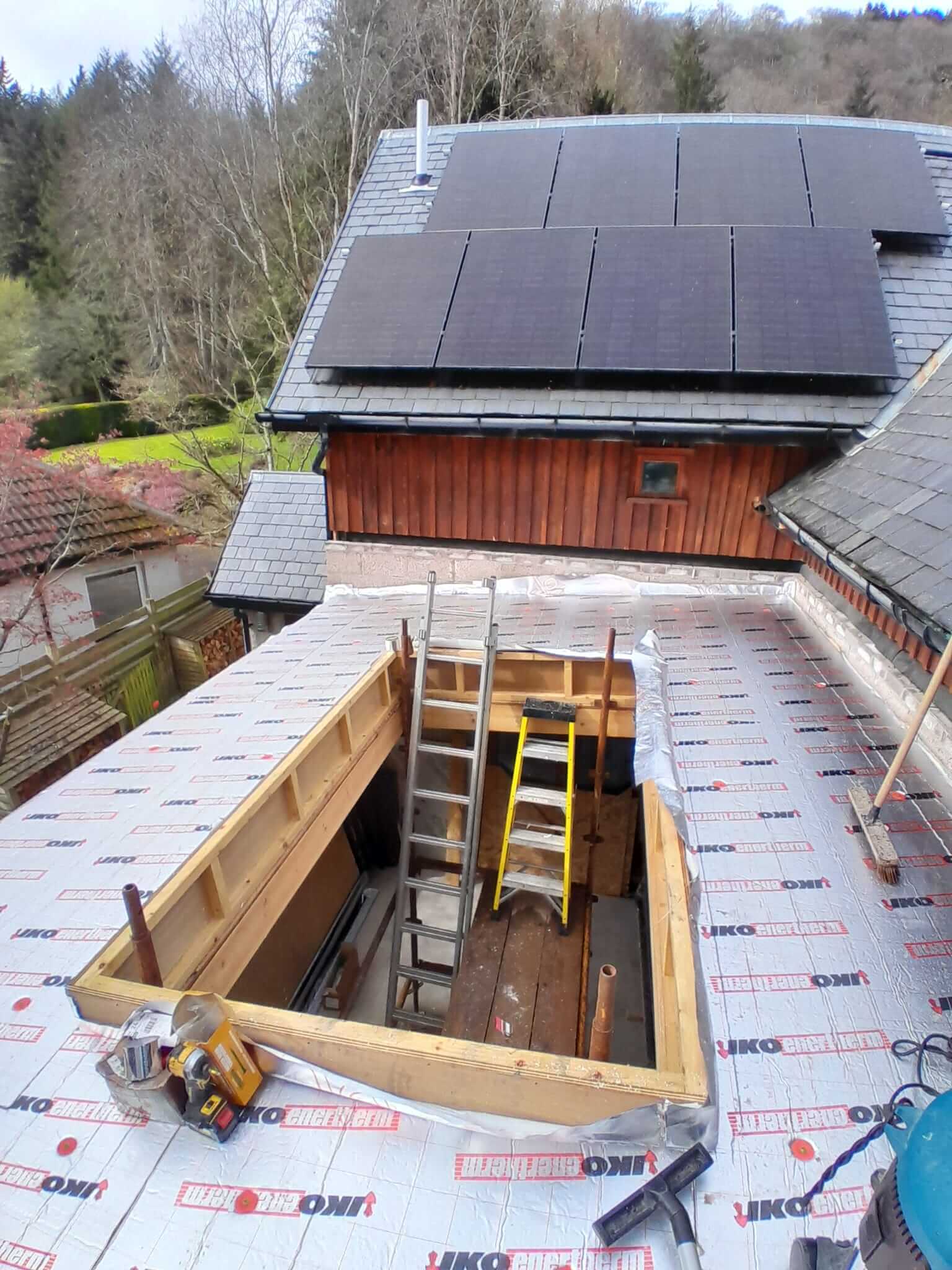 Aerial view from roof showing large rooflight opening framed out in flat roof deck with IKO membrane laid, solar panels on main house roof beyond