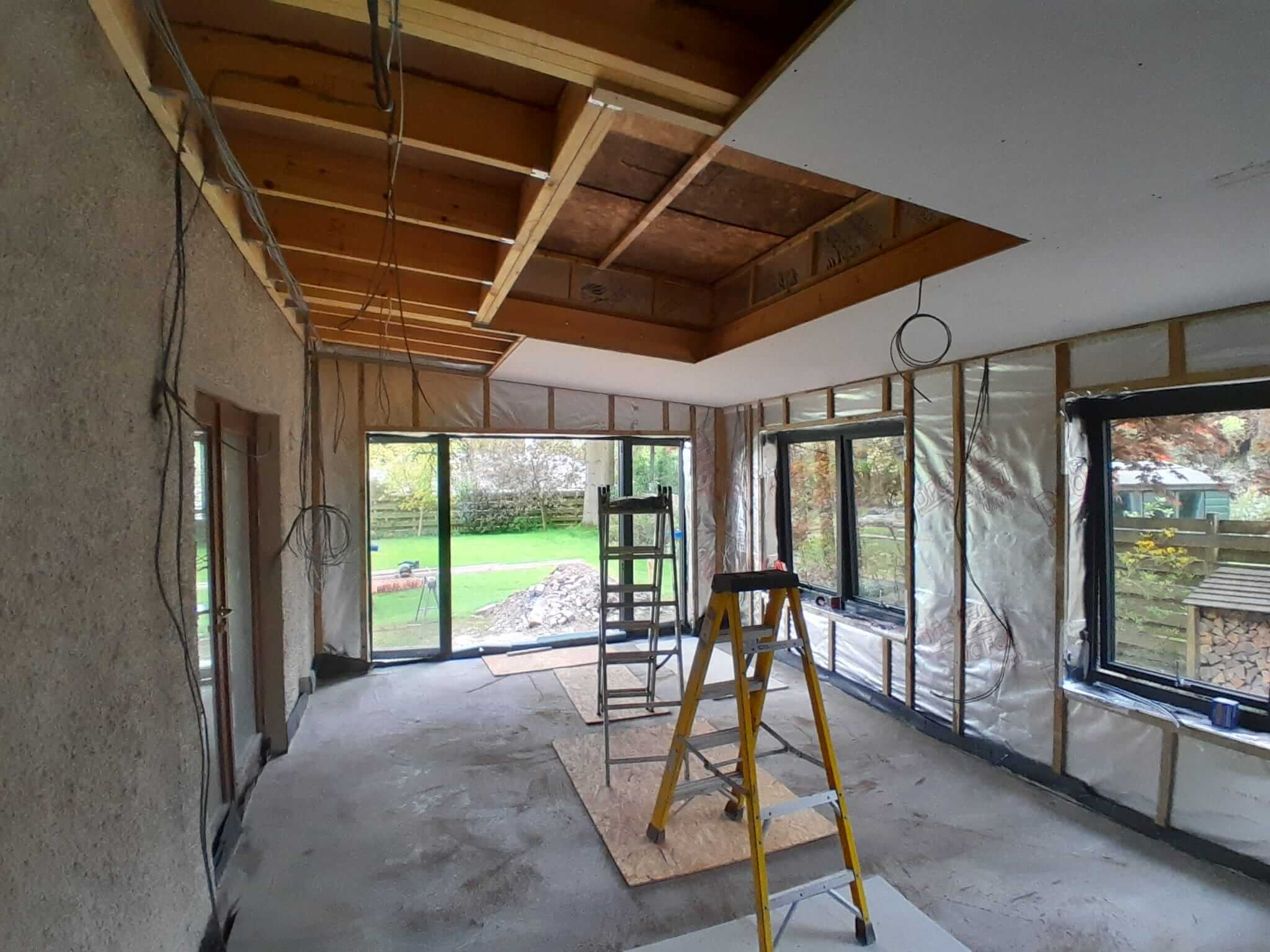 Interior of extension at first fix stage, ceiling boarding partially complete over rooflight void, bifold doors open to garden, first fix wiring and vapour barrier visible