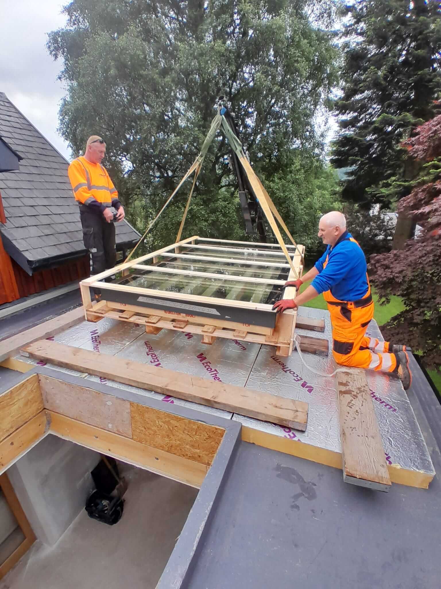 Two operatives guiding bespoke glazed rooflight unit into position on flat roof using mini crane, EcoTherm insulation boards visible