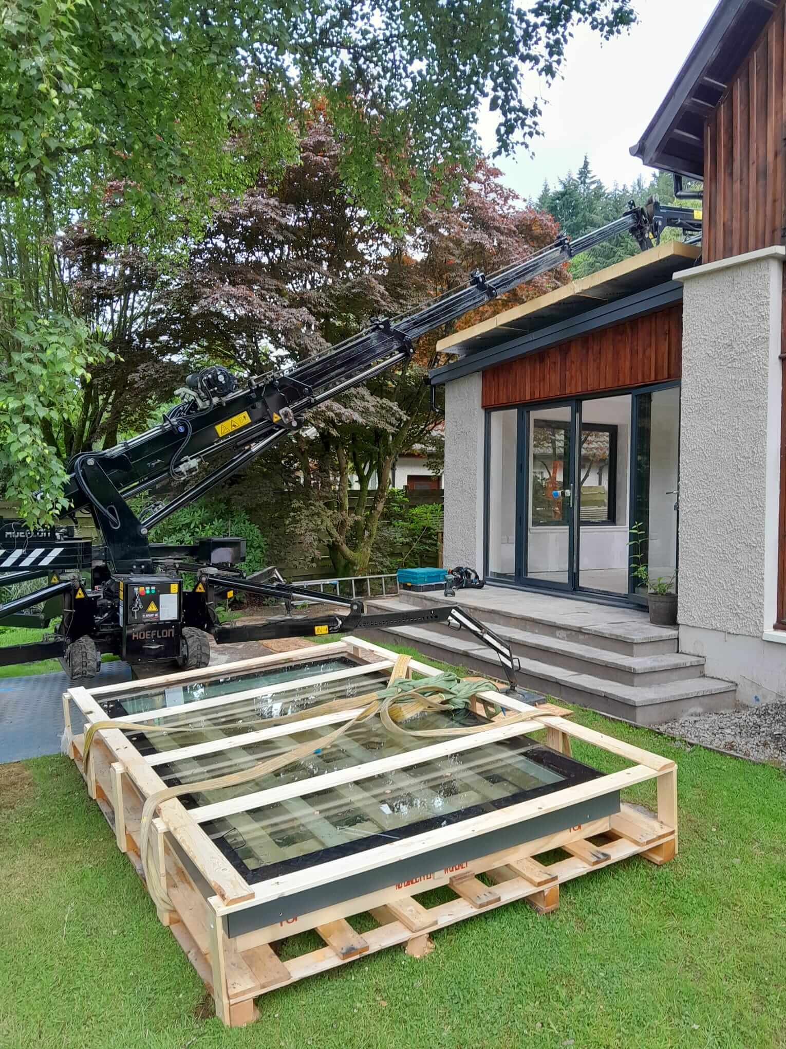 Bespoke rooflight unit in timber delivery crate on lawn, Hoeflon mini crane positioned ready to lift, completed extension entrance beyond