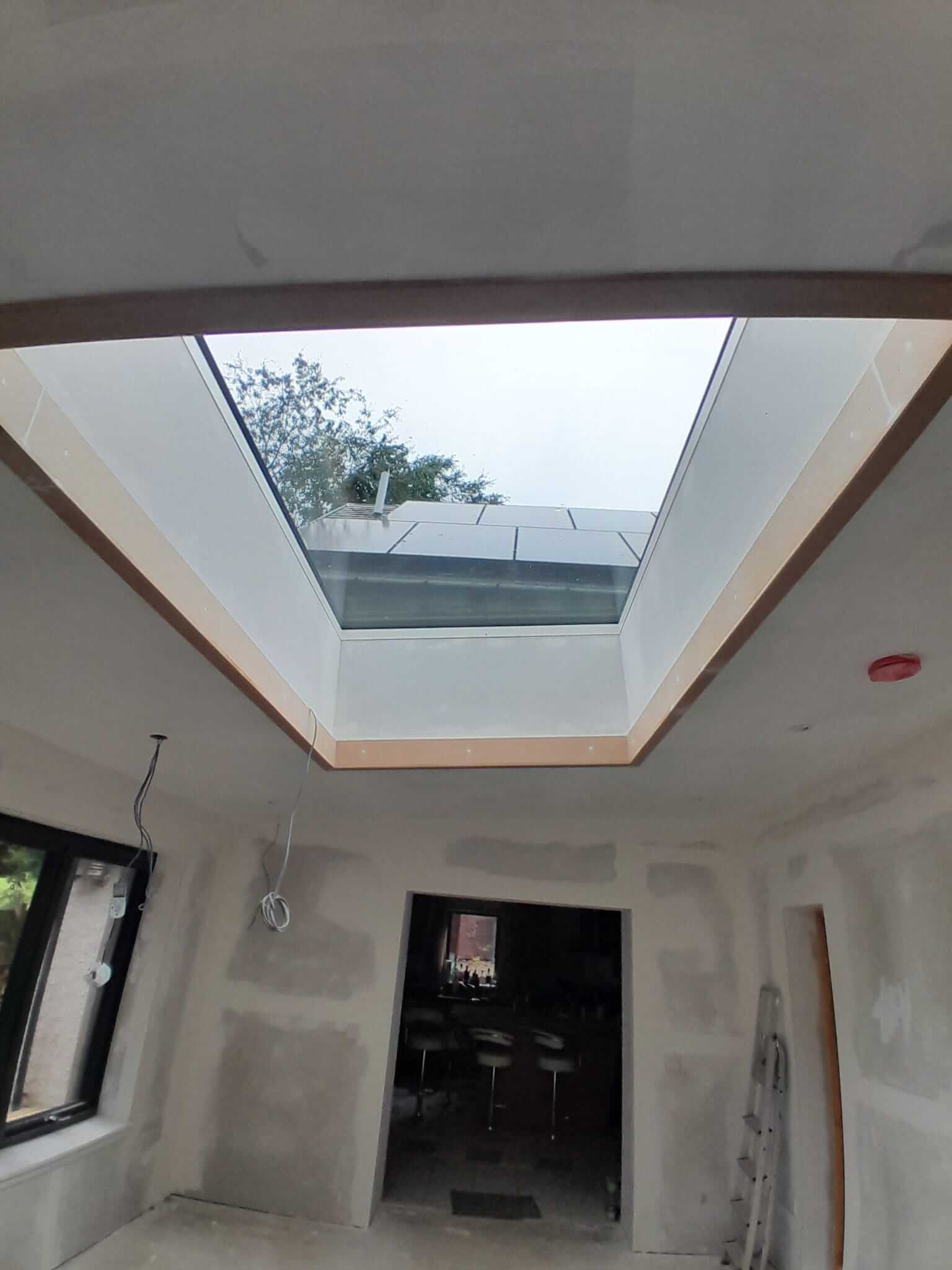 Interior view looking up at installed bespoke rooflight flooding plastered extension with natural light, oak-trimmed reveal, main house visible through doorway below