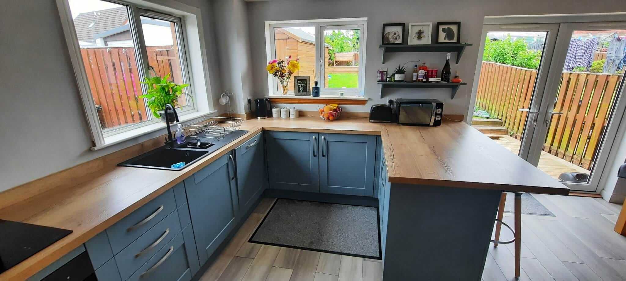 Completed open-plan kitchen with blue-grey shaker cabinets, oak worktops, floating shelves and French doors to the garden