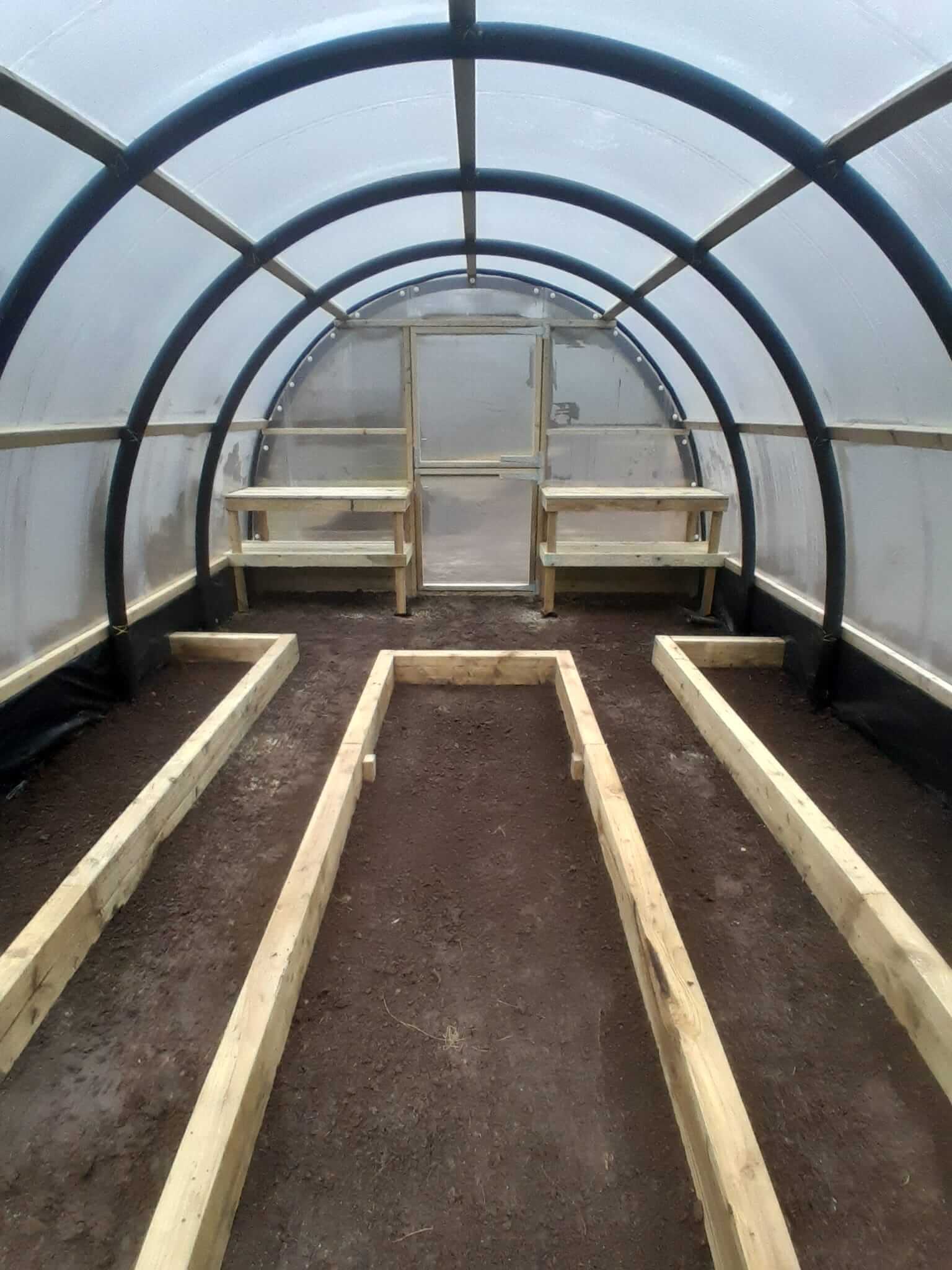 Interior of Poly Crub tunnel looking toward stable door, timber raised planter bed frames laid out on soil floor, potting benches either side